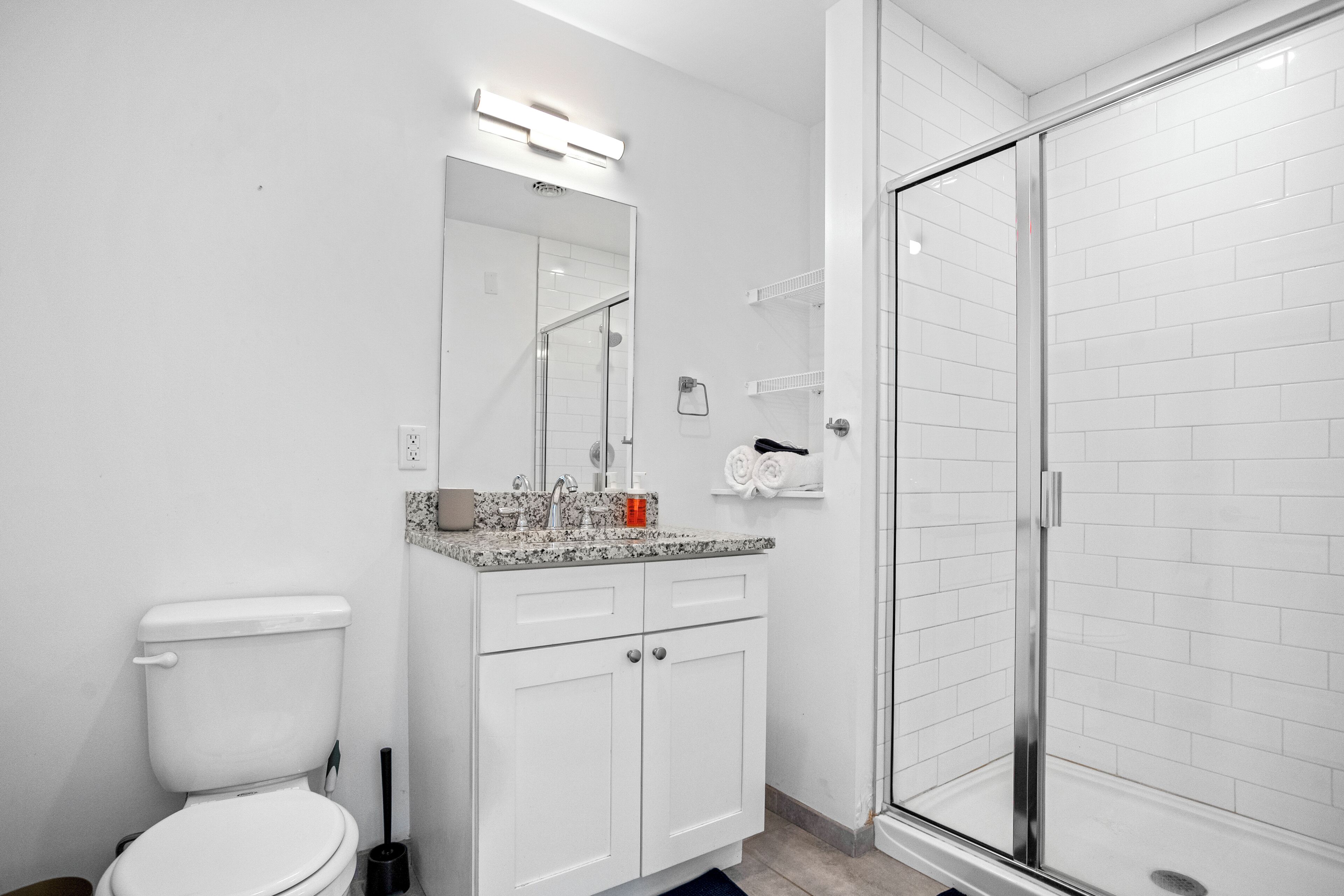 Un bany modern amb un vàter blanc, un lavabo amb taulell de granat, una cabina de dutxa de vidre i parets revestides de rajoles blanques.