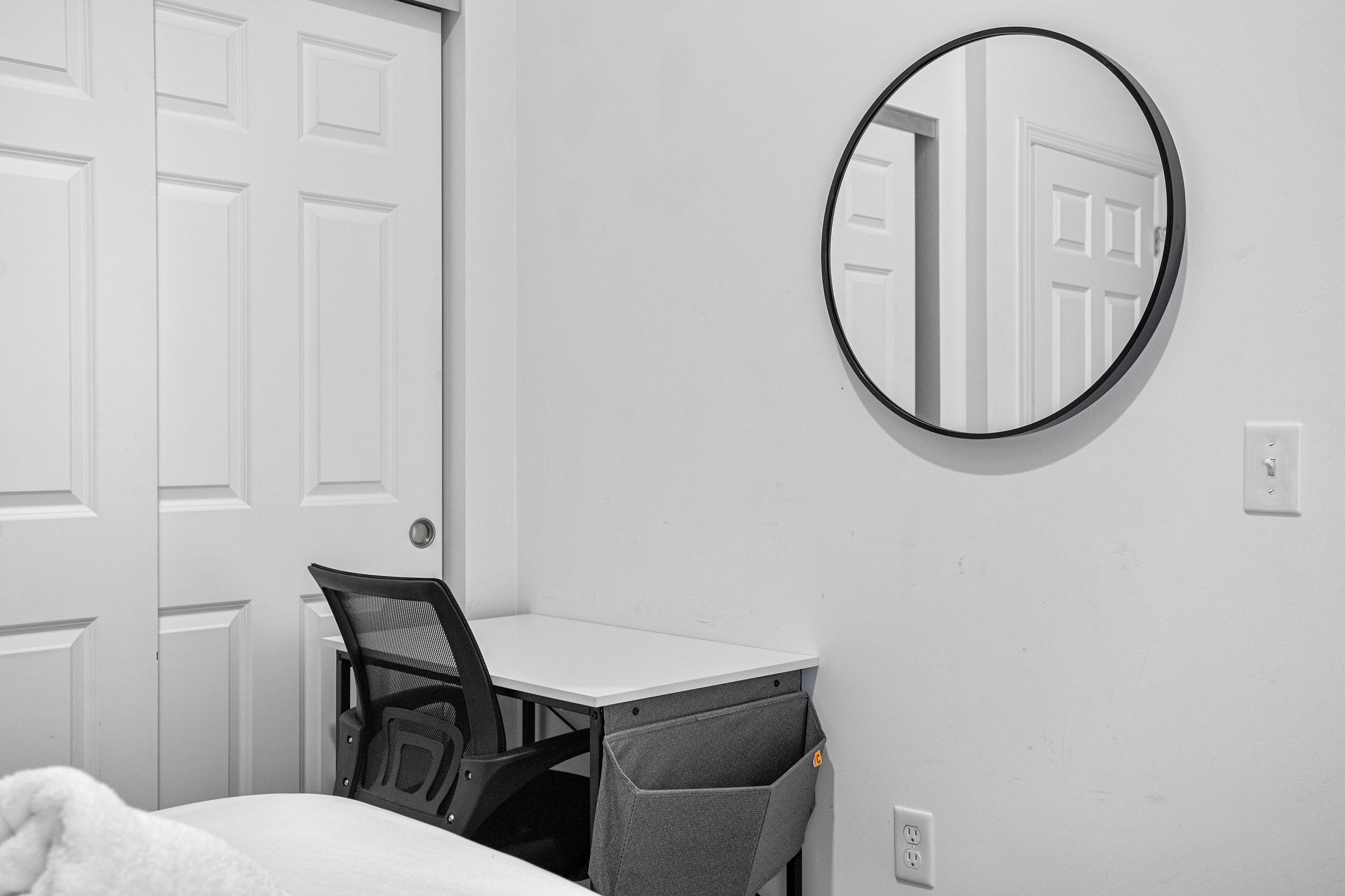 Una camera da letto con un letto bianco, una scrivania con una sedia, uno specchio rotondo e una porta bianca.