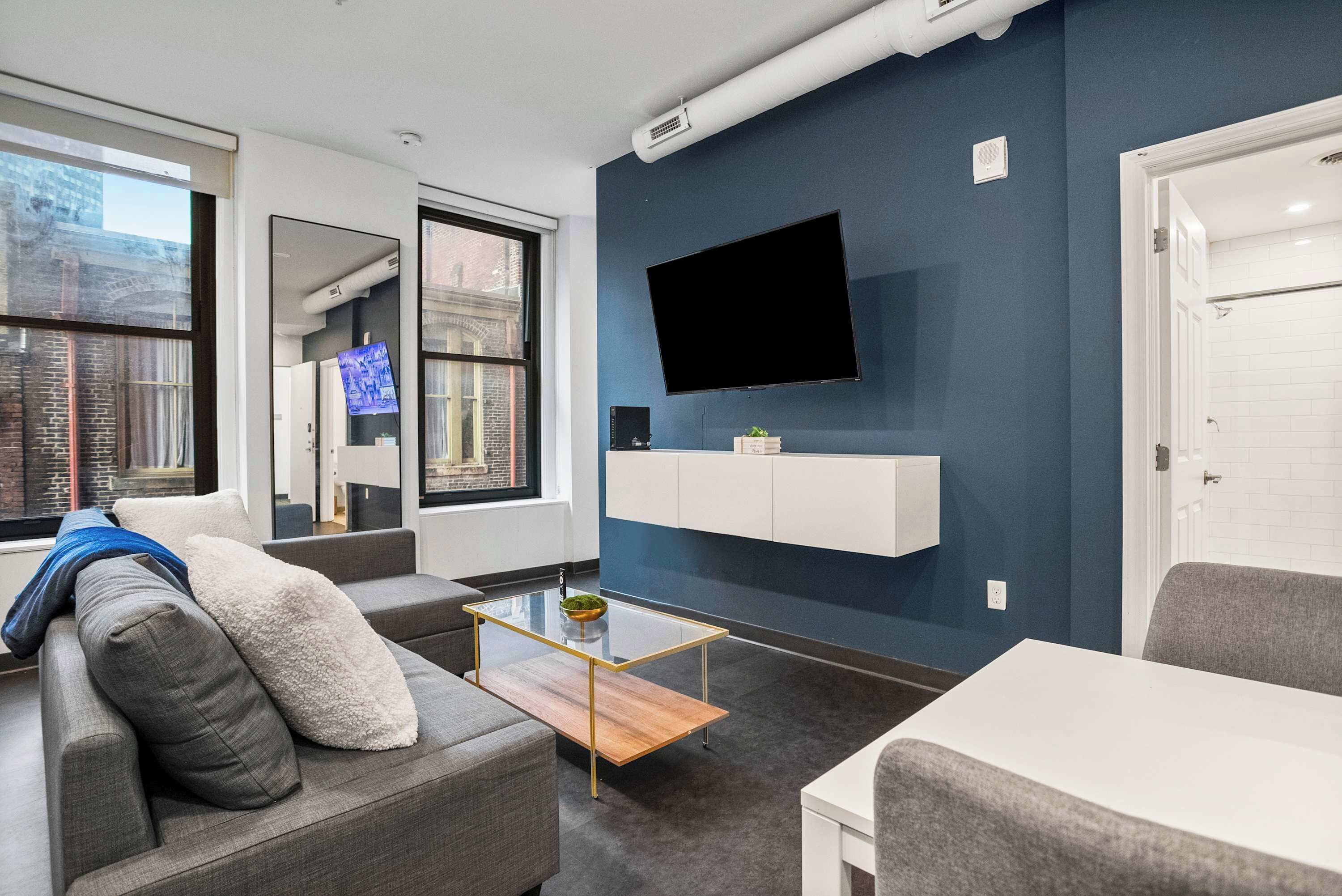 A modern living room with a grey sofa, a glass coffee table, a mounted TV, and a white cabinet against a blue wall.