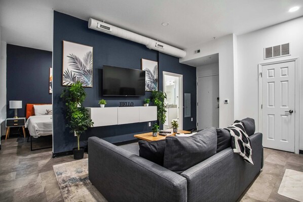 A modern living room with a grey sofa, a flat-screen TV, and two potted plants. - CozySuites - William (Pittsburgh)