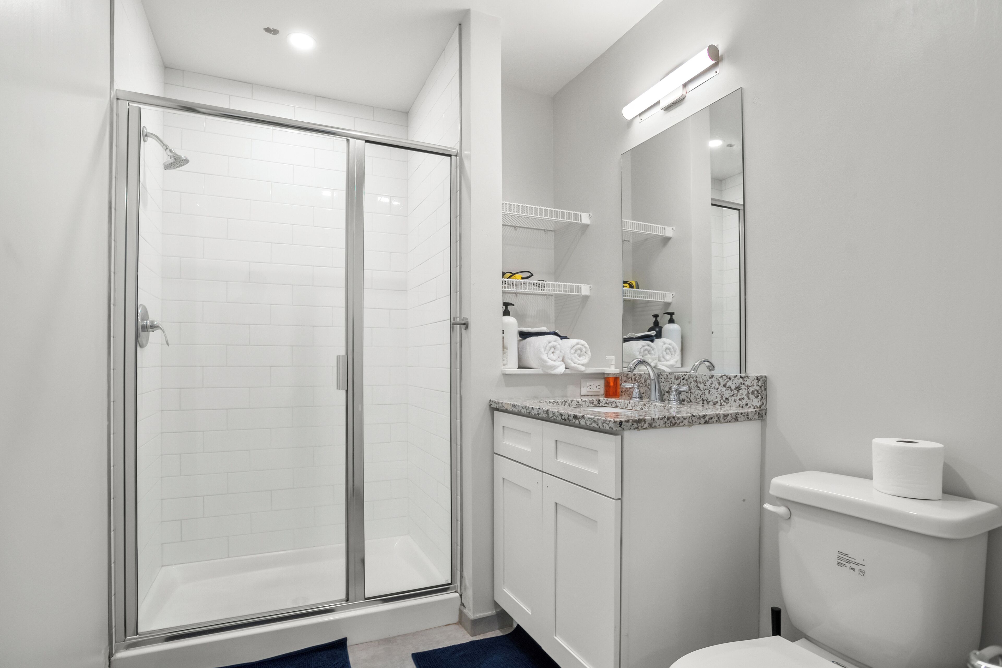 A modern bathroom with a glass-enclosed shower, white cabinetry, a marble countertop, and a toilet with a paper towel dispenser.