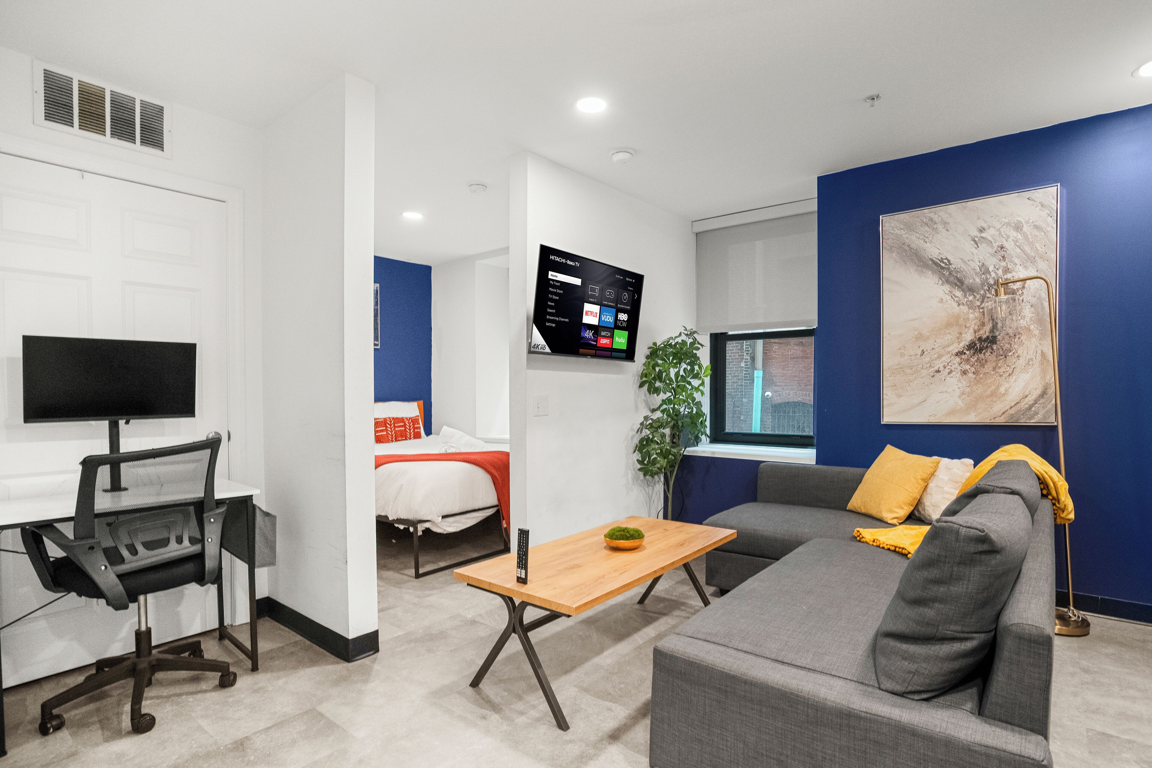 Een moderne hotelkamer met een grijze bank, een houten salontafel, een bureau met een computer, een aan de muur bevestigde televisie en een groot abstract schilderij.