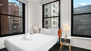 A hotel room with a large bed, two bedside tables, and large windows offering city views. - CozySuites - William (Pittsburgh)