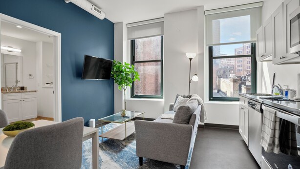 A modern kitchen with white cabinets, a built-in oven, and a large window offering a city view. - CozySuites - William (Pittsburgh)