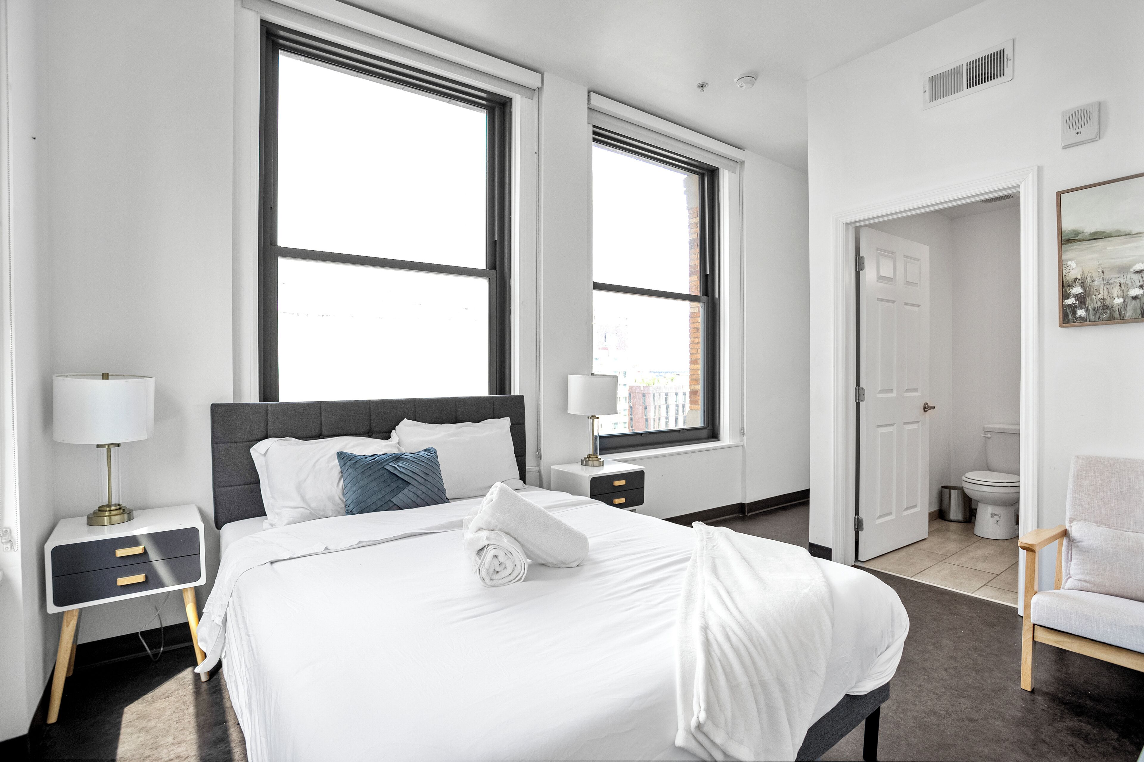 A hotel room with a large bed, two bedside tables, a chair, and a view of the outside through large windows.