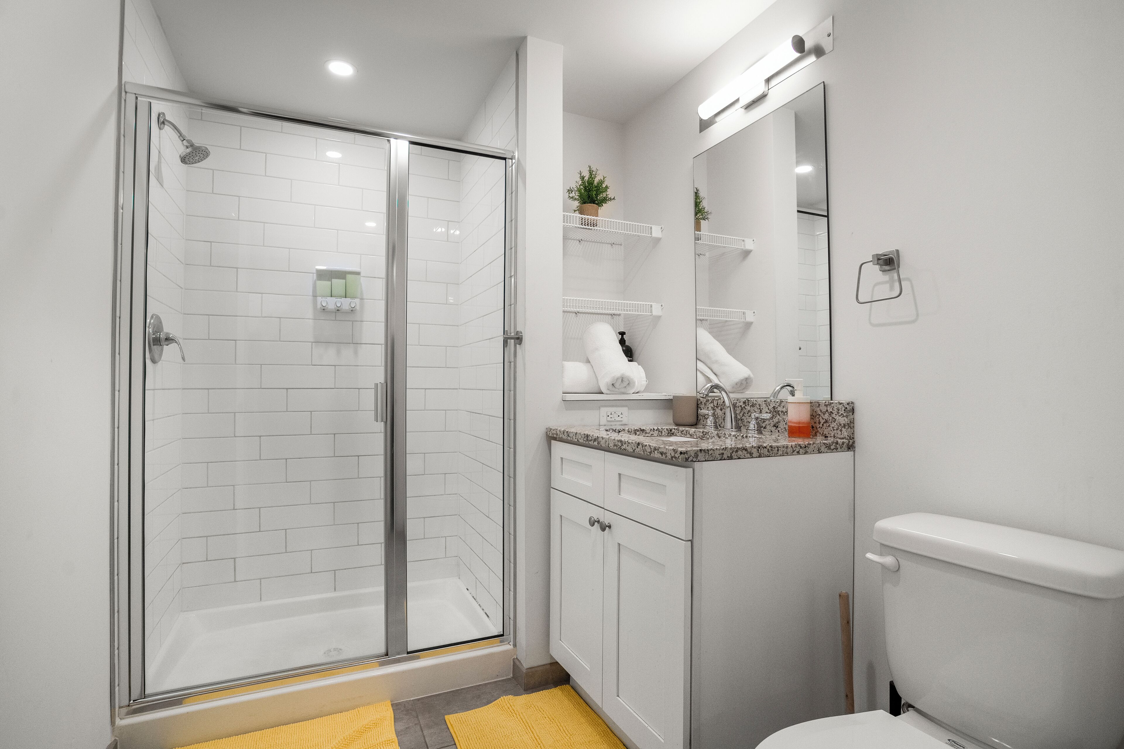 A modern bathroom with a glass-enclosed shower, white tiled walls, a granite countertop, and a toilet.