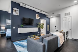 A modern living room with a grey sofa, a flat-screen TV mounted on the wall, a small coffee table, and a blue rug. - CozySuites - William (Pittsburgh)