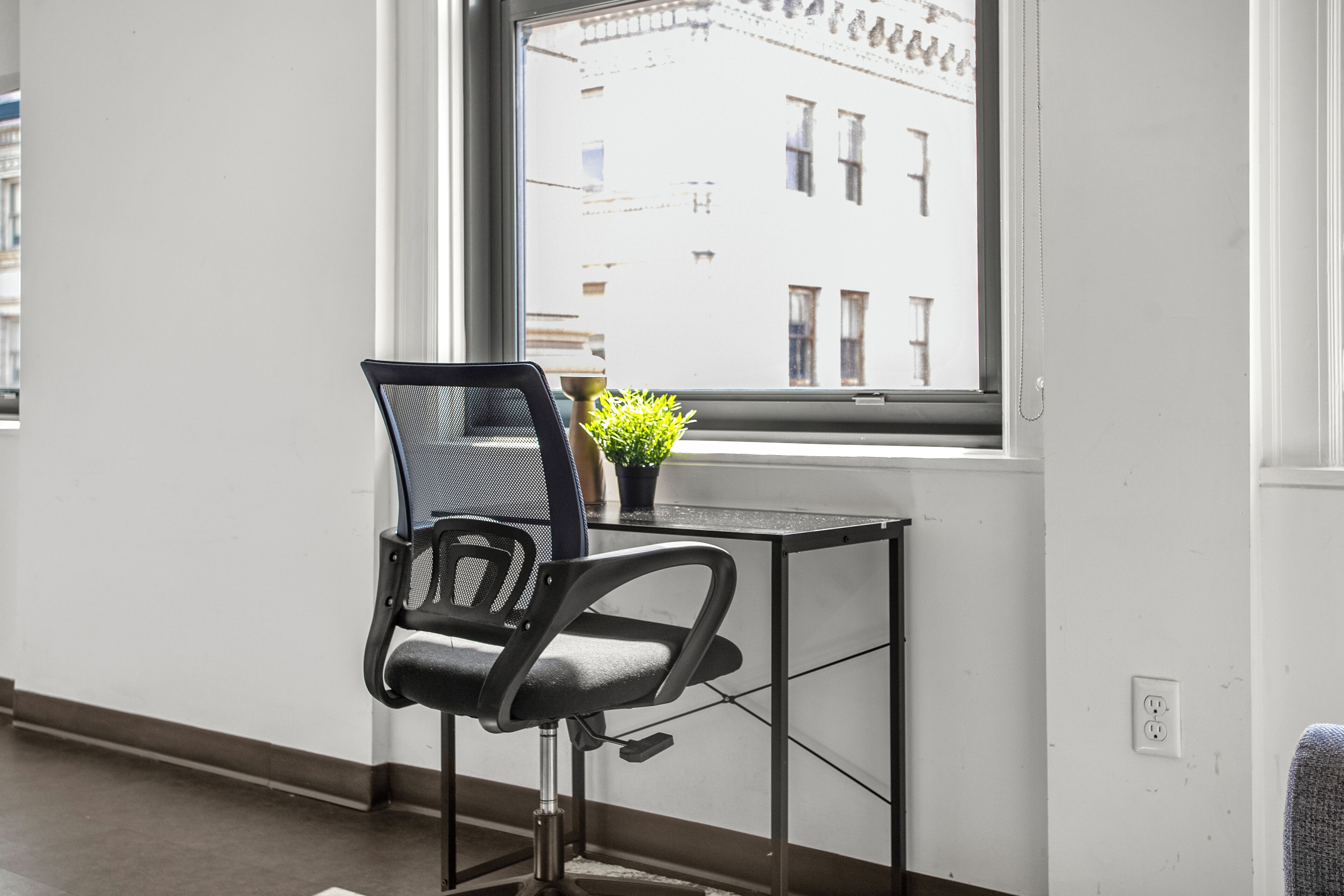 Une chaise de bureau moderne, dotée d’un dossier en tissu à mailles et d’une structure en métal, est placée à côté d’une petite table sur laquelle se trouve un pot de fleurs. On aperçoit, au fond, une fenêtre donnant sur un immeuble.