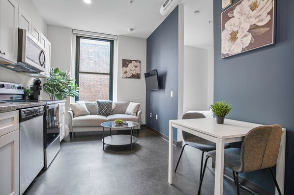 A modern kitchen with white cabinets, a dining area with a white table and chairs, a gray sofa, and a large window with a view of a brick building.