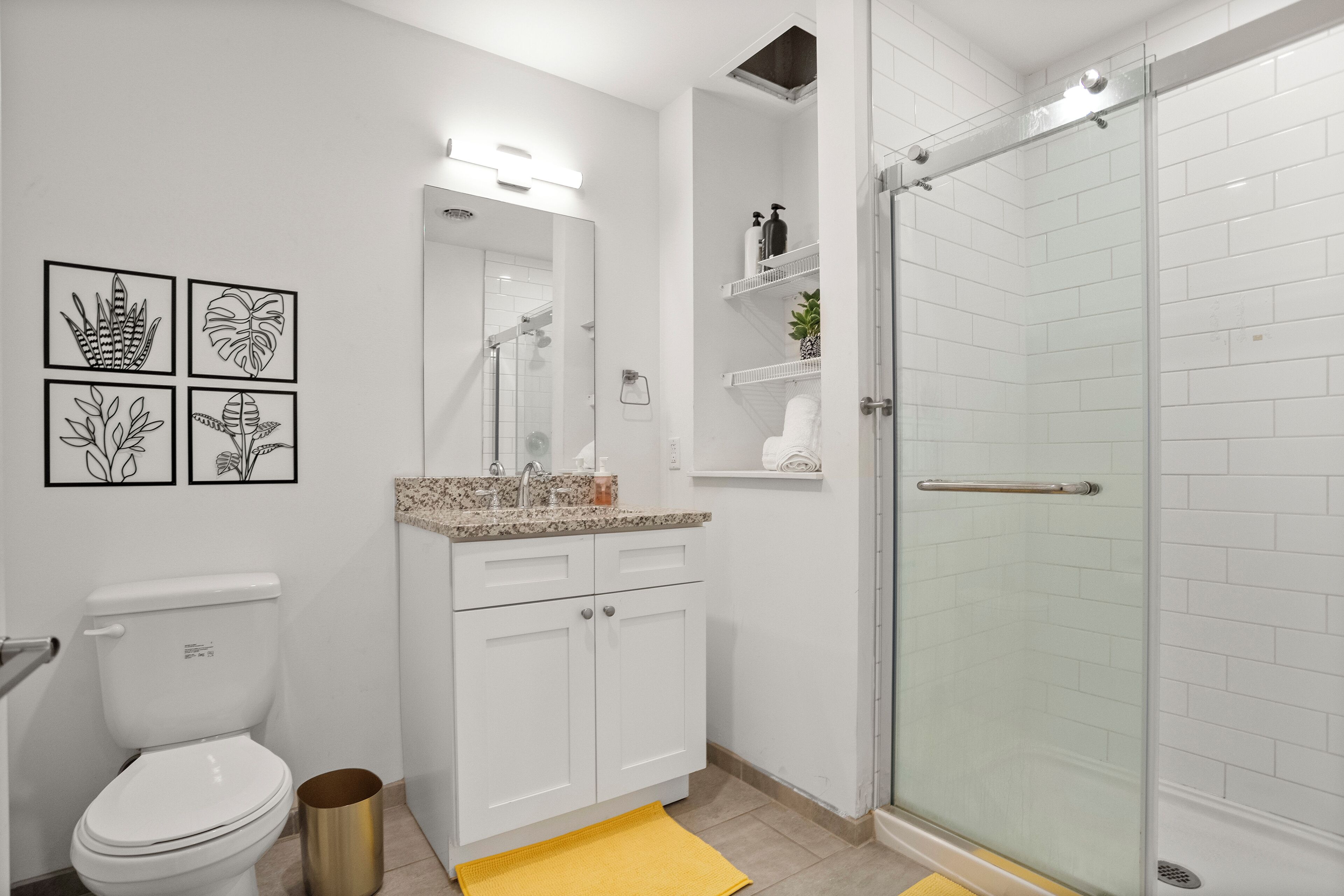Une salle de bain moderne avec des toilettes, un lavabo et une douche. Les murs sont ornés d’illustrations botaniques encadrées, et un tapis de bain jaune est posé au sol.