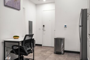 Eine kleine Küche mit einem weißen Tisch, einem schwarzen Bürostuhl, einer Edelstahl-Mülltonne und einem Kühlschrank.