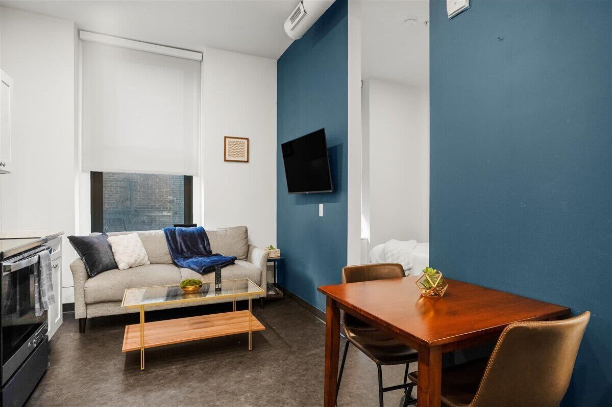 A modern living room with a sofa, coffee table, and dining table. A flat-screen TV is mounted on the wall.