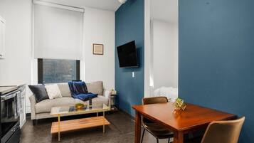 A modern living room with a sofa, coffee table, and dining table. A flat-screen TV is mounted on the wall.