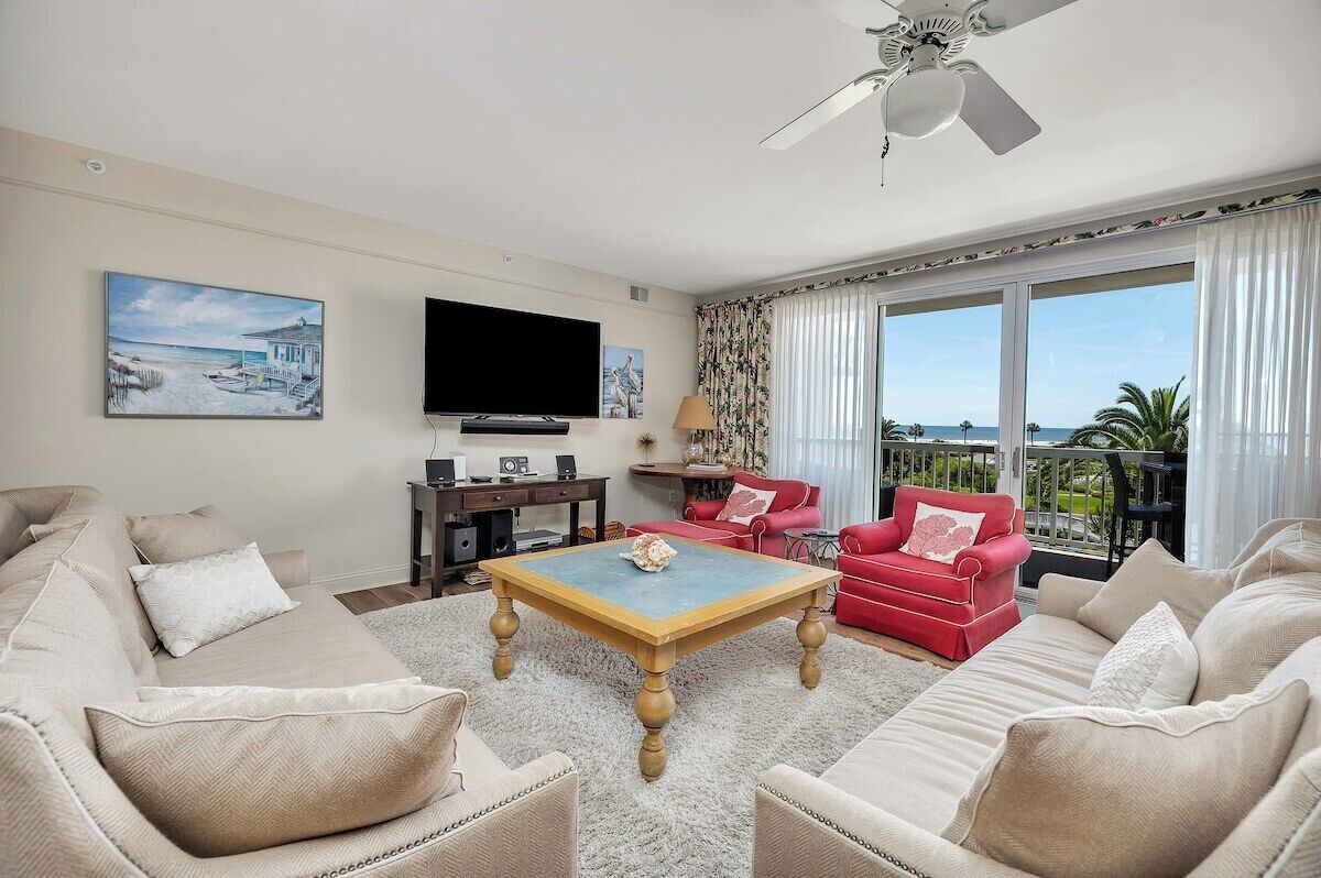 A living room with a beige sofa, a red armchair, a wooden coffee table, a flat-screen TV, and a balcony with a view of the ocean.