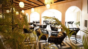 Un restaurant doté d’une terrasse, avec des chaises et des tables noires, entouré d’arcades et de plantes.