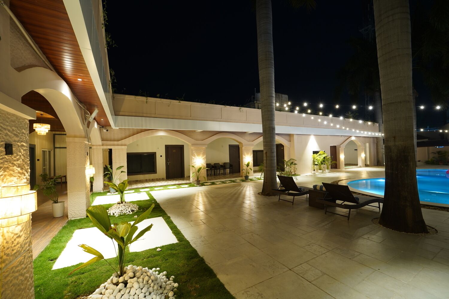 A well-lit outdoor area with a swimming pool, lounge chairs, and a patio with a stone wall and greenery.