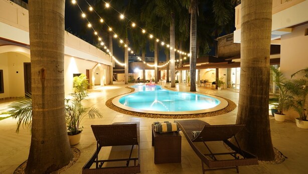 Un espace piscine doté de chaises longues et illuminé de guirlandes lumineuses, entouré de palmiers et bordé d’un bâtiment à plusieurs étages.