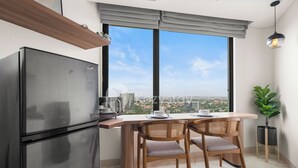 A compact kitchen with a modern refrigerator, a dining table with two chairs, and a large window offering a city view. - Tenui by Kozystay - Tanjung Barat (Jakarta)