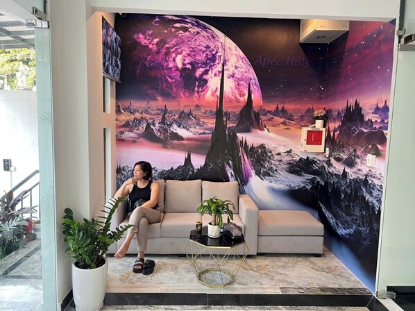 A woman sitting on a beige sofa in a lobby with a large space-themed mural, a potted plant, and a small table with a vase. - HOA PHU Hotel (Ho Chi Minh City)