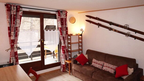 Una sala de estar acogedora con un sofá marrón, cojines rojos, una mesa de madera y una ventana con cortinas.