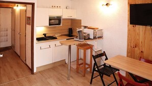 Una cocina compacta con microondas, tostadora y hervidor. Hay una mesa de madera con dos sillas y un taburete. La habitación tiene paredes blancas y suelo de madera.