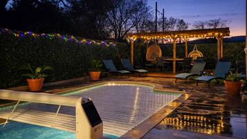 Una piscina exterior con un muelle de madera, sillas de descanso y una pérgola con luces de cuerda.