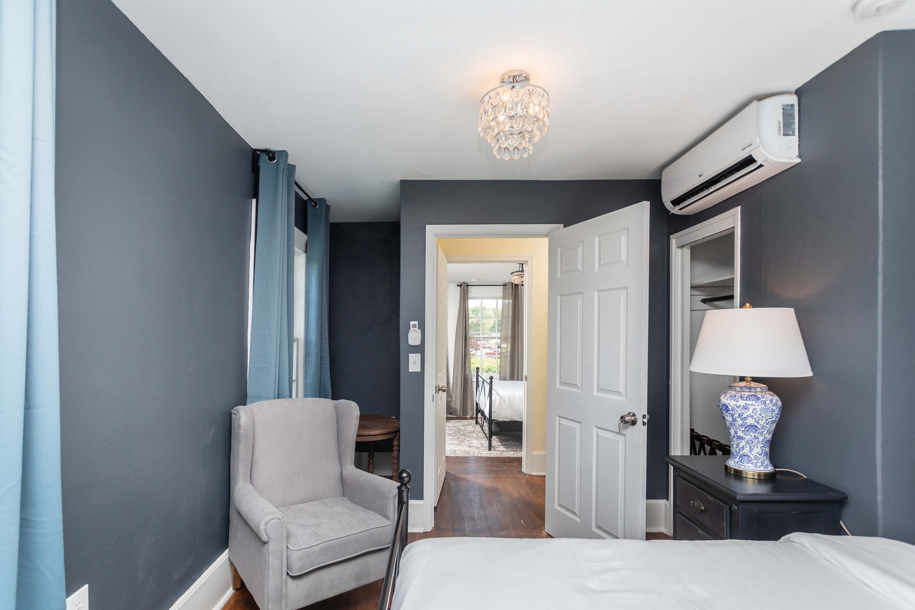 A hotel room with a bed, a chair, a nightstand with a lamp, an air conditioning unit, and a chandelier.