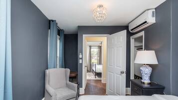 A hotel room with a bed, a chair, a nightstand with a lamp, an air conditioning unit, and a chandelier.