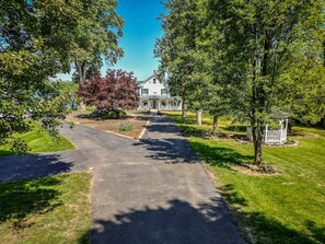 A residential driveway leading to a house with a porch, surrounded by trees and a well-maintained lawn.