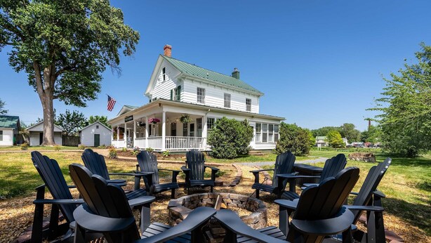A white house with a green roof, a porch with chairs, and a fire pit with chairs around it. - Charming suite w/ TV & patio (Luray)