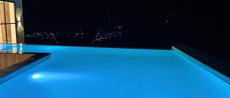 Piscina infinita con terraza de madera y vista nocturna de la ciudad.