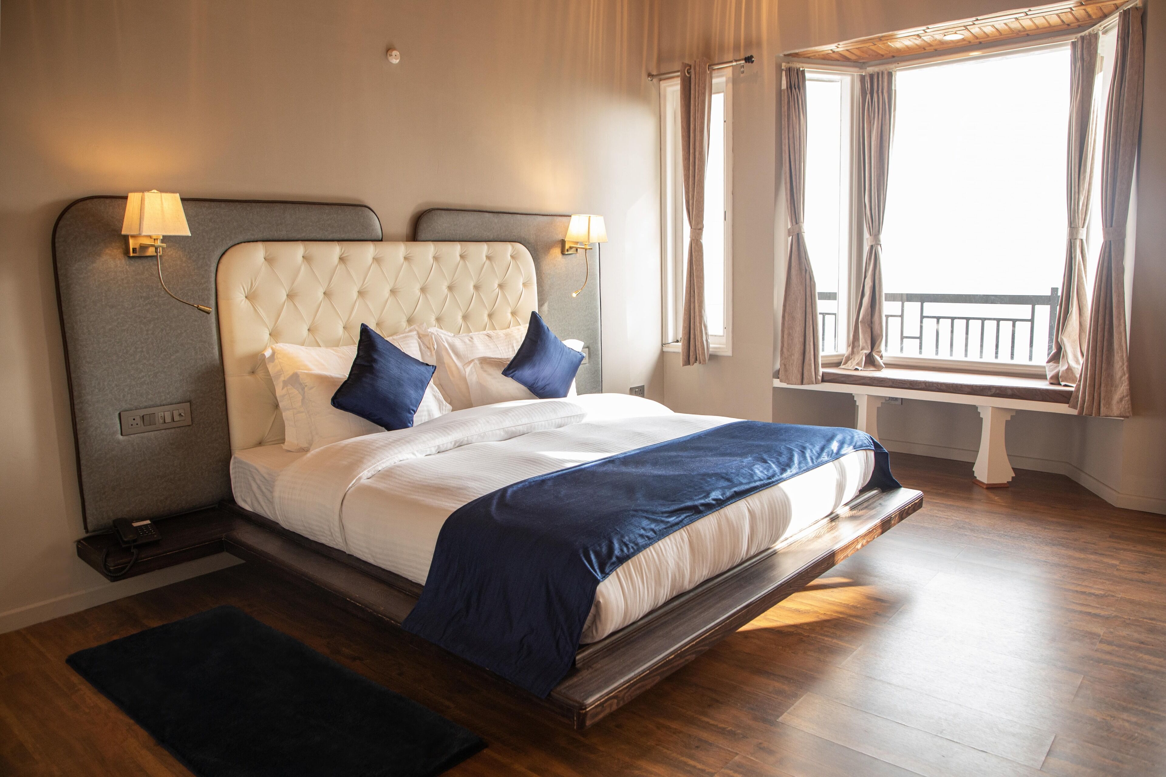 Een keurig opgemaakt bed met een gestoffeerd hoofdbord, twee bedlampjes en uitzicht op het balkon door een raam met gordijnen.