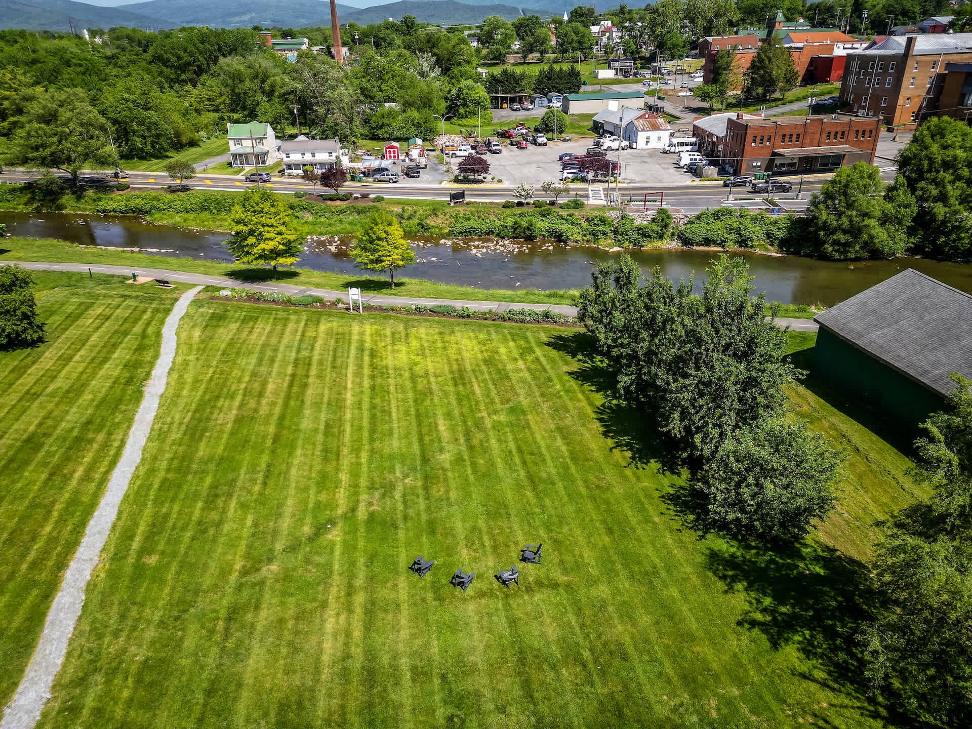 A large green field with a paved path, surrounded by buildings and a body of water.