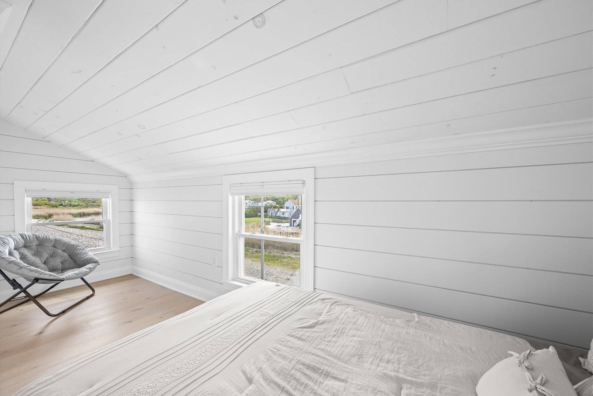 Un dormitorio con una cama, una silla y dos ventanas que dan a un espacio verde y casas.