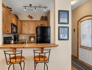 A kitchen with wooden cabinets, a black refrigerator, a coffee maker, and two bar stools. - Sun Peaks 67 | Steps from the Village (Sun Peaks)