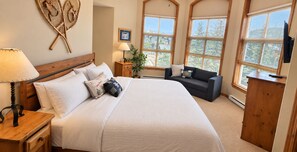 A bedroom with a large bed, a nightstand, a lamp, a dresser, a window with a view, and a wall-mounted decoration. - Sun Peaks 67 | Steps from the Village (Sun Peaks)