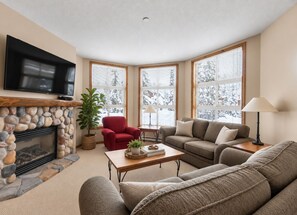 A cozy living room with a stone fireplace, a red armchair, a grey sofa, and a television mounted on the wall. - Sun Peaks 67 | Steps from the Village (Sun Peaks)