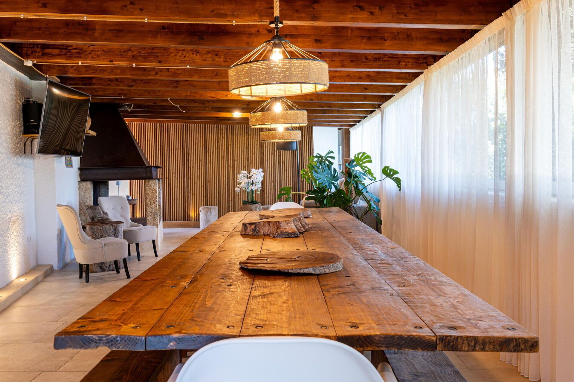 Una larga mesa de comedor de madera con diseño rústico, rodeada de sillas blancas, una lámpara colgante y un muro de madera con listones verticales.
