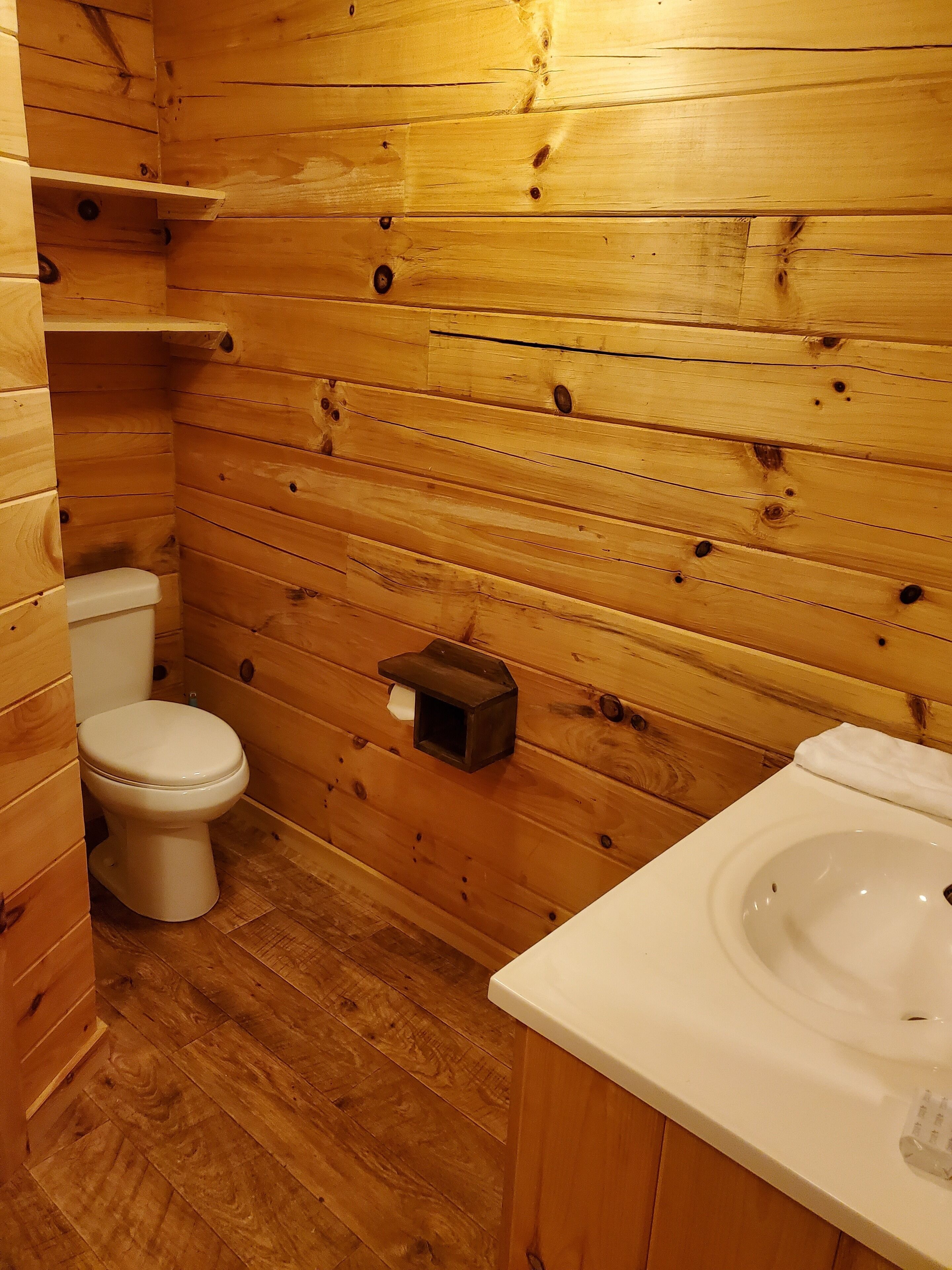 A wooden-paneled bathroom with a toilet, a sink, and a small shelf.