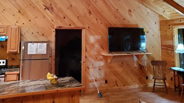 Interior acogedor de una cabaña con techo de madera, ventilador de techo, zona de cocina con refrigerador y estufa, comedor con mesa y sillas, y sala de estar con sofá.