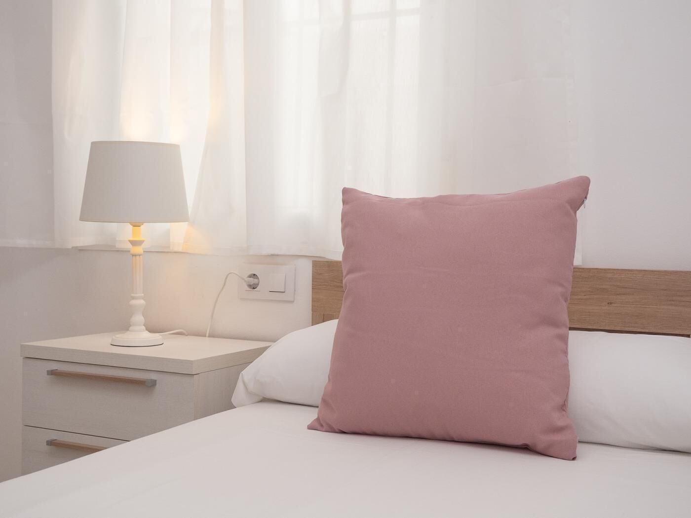 A neatly made bed with a single pink pillow and a white bedside table with a lamp.