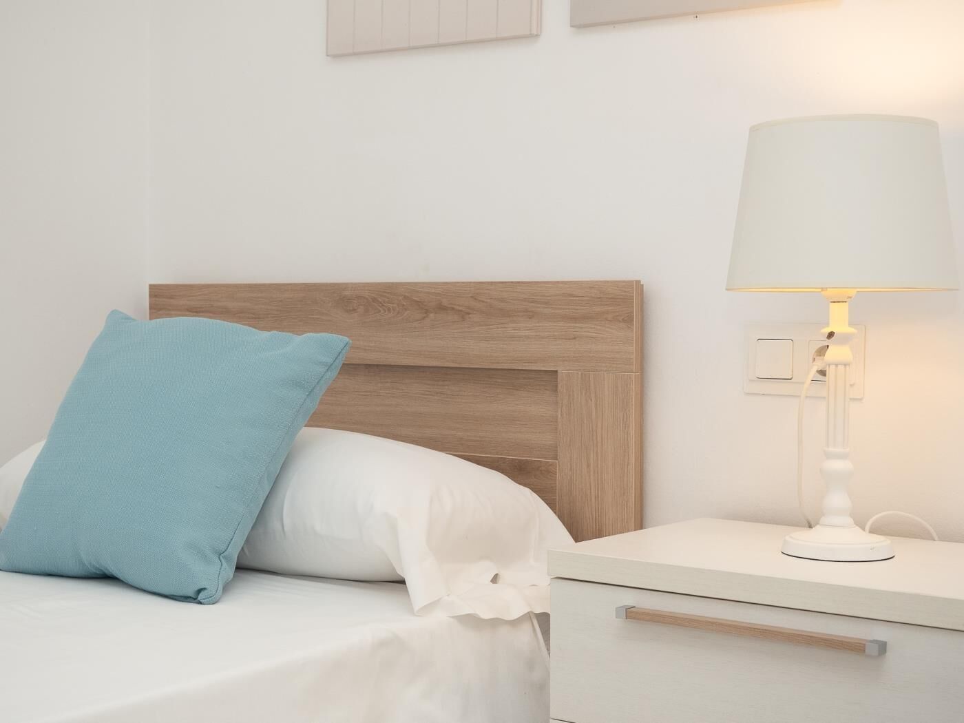 A neatly made bed with a wooden headboard, a white nightstand with a lamp, and a light switch on the wall.