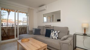 A modern living room with a beige sofa, a wooden coffee table, a glass side table with a lamp, and a large window with a view of buildings and trees. - sibssanlúcar Camelias (Sanlucar de Barrameda)