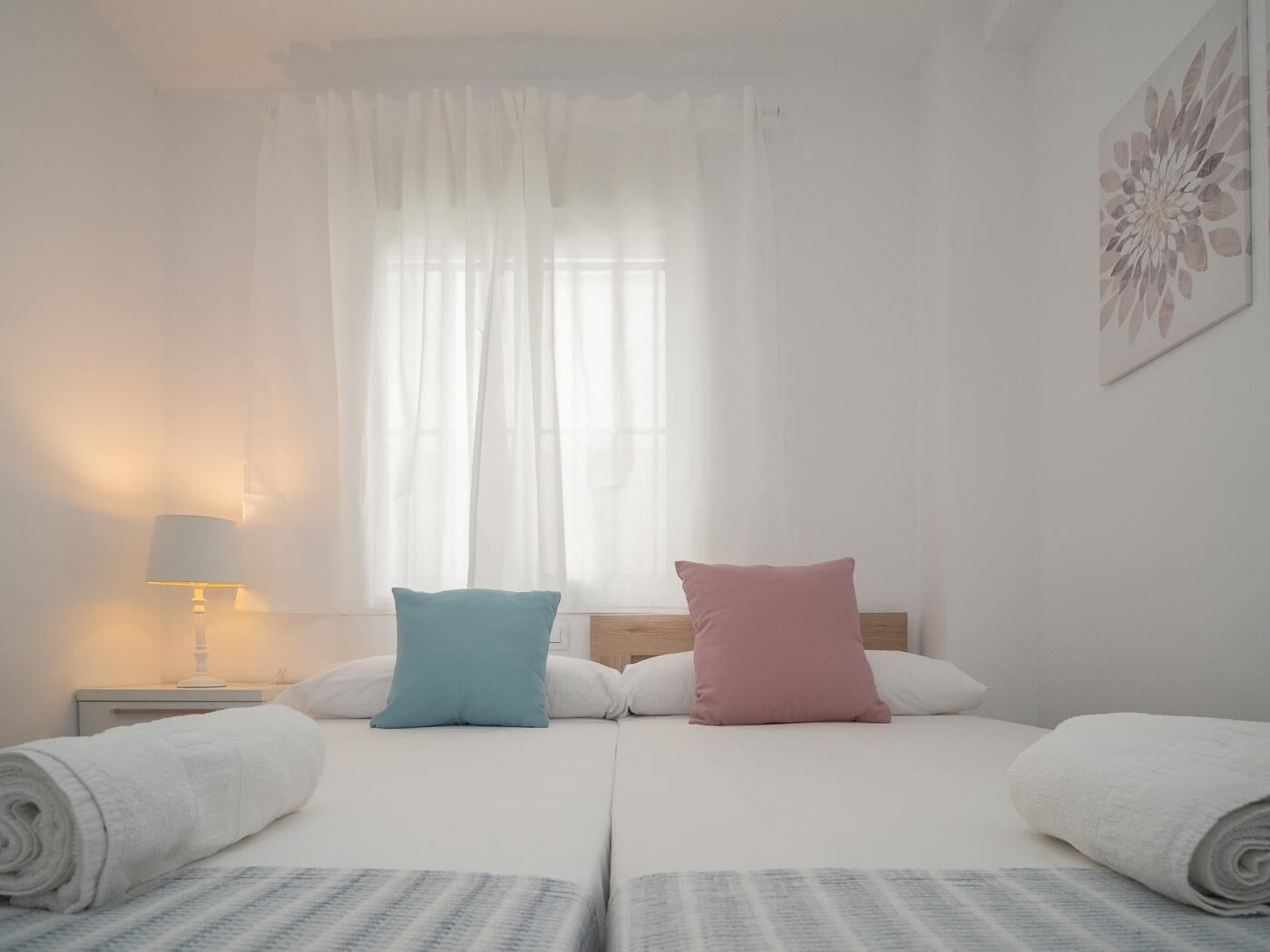 A hotel room with two beds, a nightstand with a lamp, a window with sheer curtains, and a wall-mounted artwork.