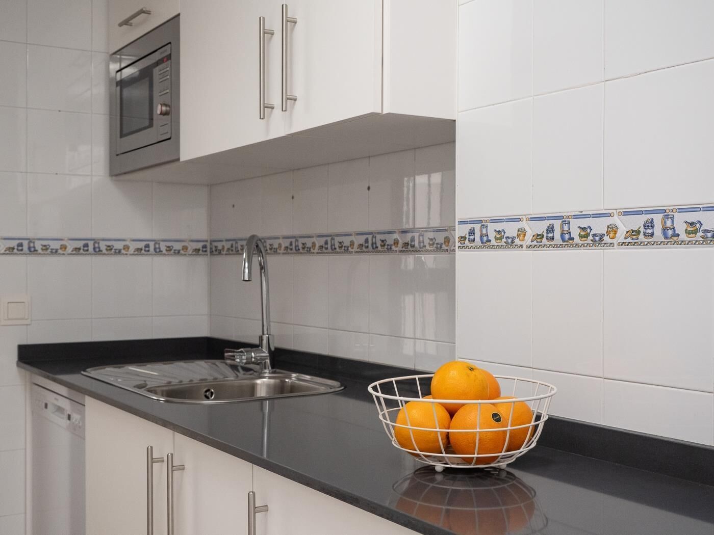 Een keuken met witte kastjes, een roestvrijstalen spoelbak en een mand met sinaasappels.