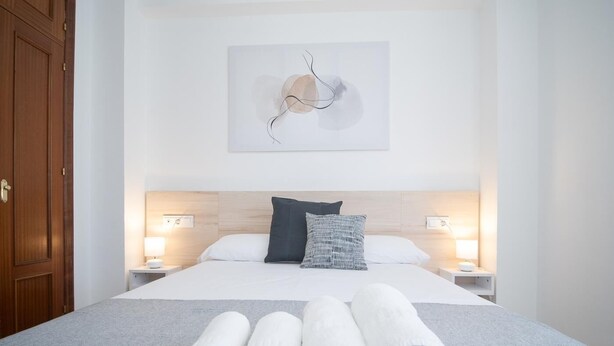 A neatly made bed with white linens and rolled towels, a wooden headboard, and a wall-mounted abstract art piece. - sibssanlúcar Camelias (Sanlucar de Barrameda)