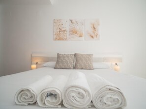 Un lit bien fait, avec des serviettes blanches pliées en rouleau, une tête de lit en bois et deux lampes fixées au mur.