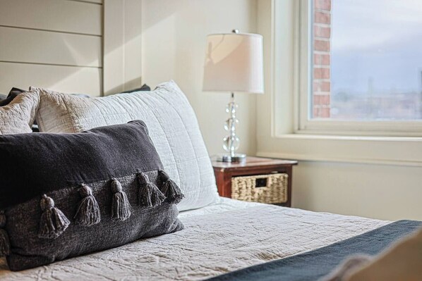 A neatly made bed with a white quilt and two pillows, one with tassels, a lamp on a bedside table, and a window with a view.