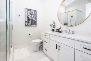 Un baño moderno con cabina de ducha de vidrio, un inodoro, un lavamanos blanco con espejo y un cuadro enmarcado en la pared.