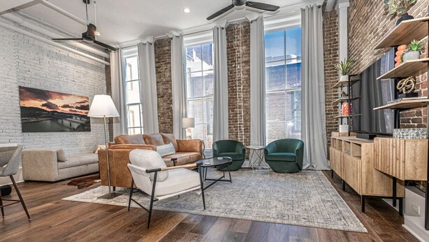 A modern living room with a brick wall, large windows, a sofa, two armchairs, a coffee table, a TV, and a bookshelf. - Downtown Luxury Condo | Near UT (Knoxville)